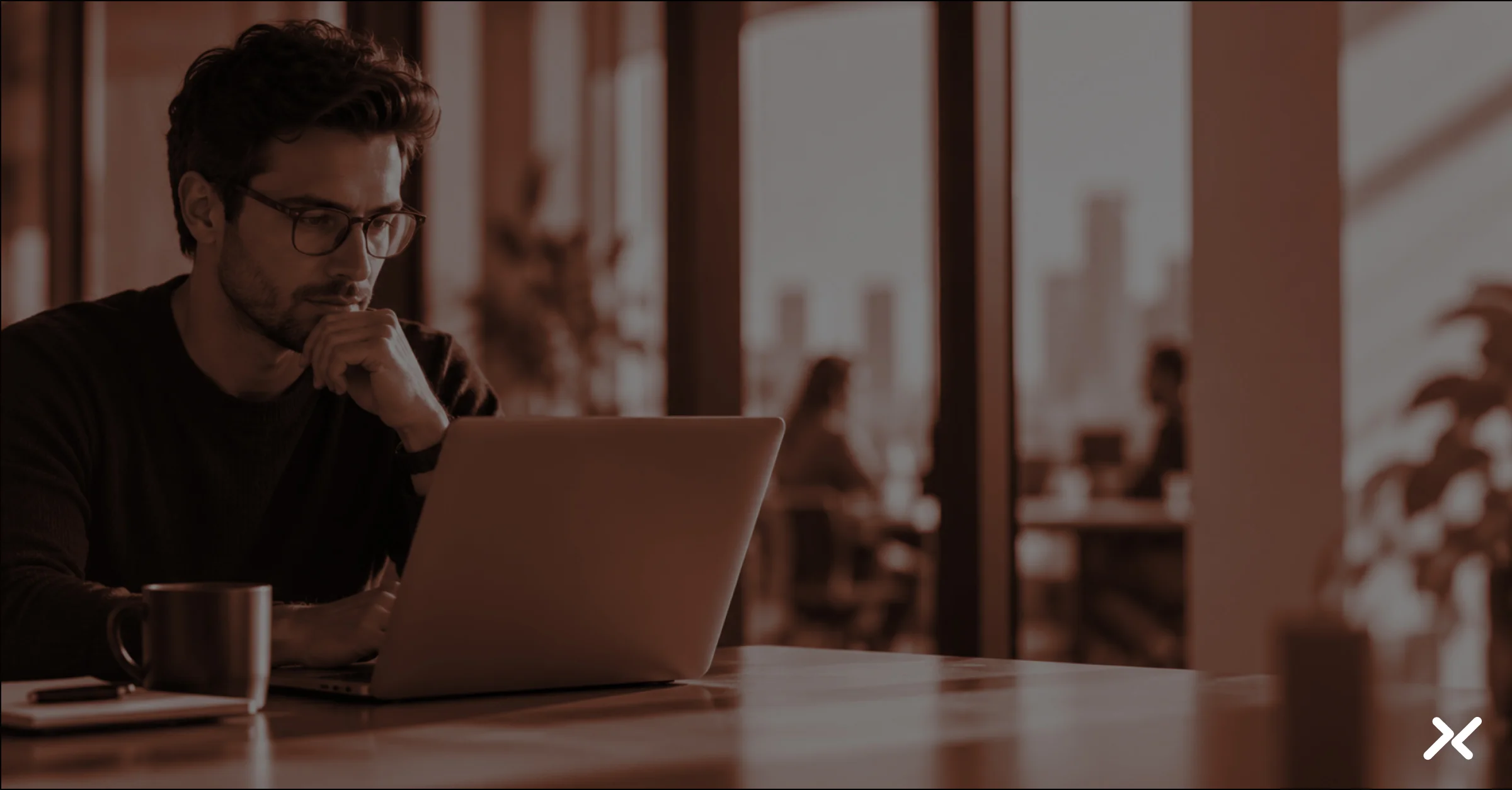 Thoughtful man in glasses works on a laptop at a sunlit office table, coffee nearby, with blurred coworkers and city skyline behind him.